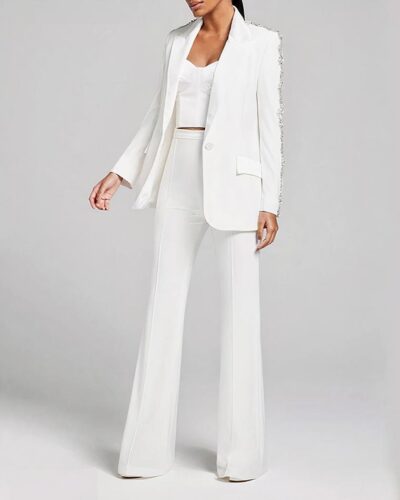 Photo d'une femme portant un ensemble blazer pantalon blanc flare avec ornements en strass sur les épaules et les manches, un top blanc le tout sur un fond gris clair