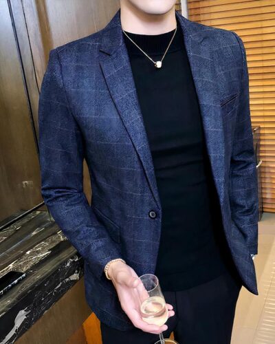 Un homme porte un blazer à carreaux bleu et un pull noir. Il a un verre à la main