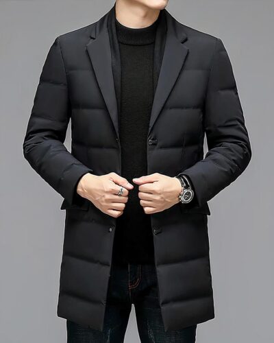 Photo d'un homme portant un blazer manteau matelassé à col amovible et boutons pression, rabats de poches et col cranté les mains tenant chacune un bord, sur un pull noir à col montant et un jean bleu foncé, le tout sur un fond gris