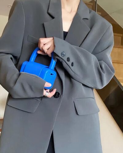 Photo d'une femme debout dans un hall devant un escalier portant un blazer oversize gris à un bouton et revers d'encolure longs, deux rabats de poche et boutons au bas des manches la main droite dans lapoche du pantalon noir et la gauche tenant un téléphone