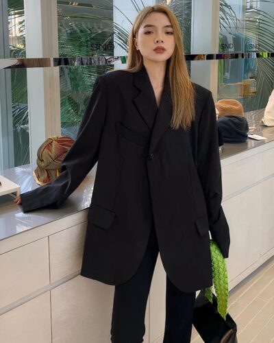 Photo d'une femm echic debout dans une boutique au grand meuble blanc, elle porte un blazer oversize noir croisé à un bouton haut sur un pantalon noir et un sac à main noir à anse verte