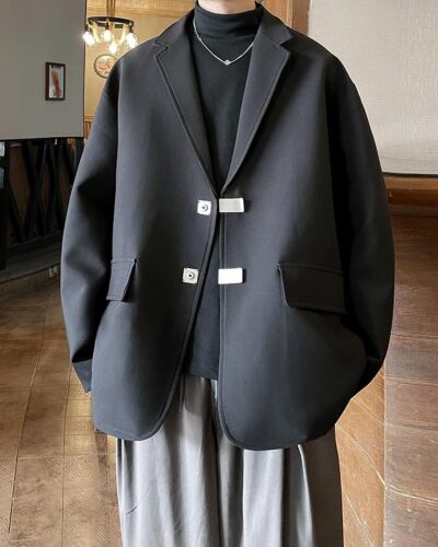 Photo d'un homme portant un blazer oversize noir à revers et grands rabats de poches factices avec deux boutons argentés carrés sur un pull fin noir col roulé et un pantalon ample gris, dans une pièce au sol marron et murs beige