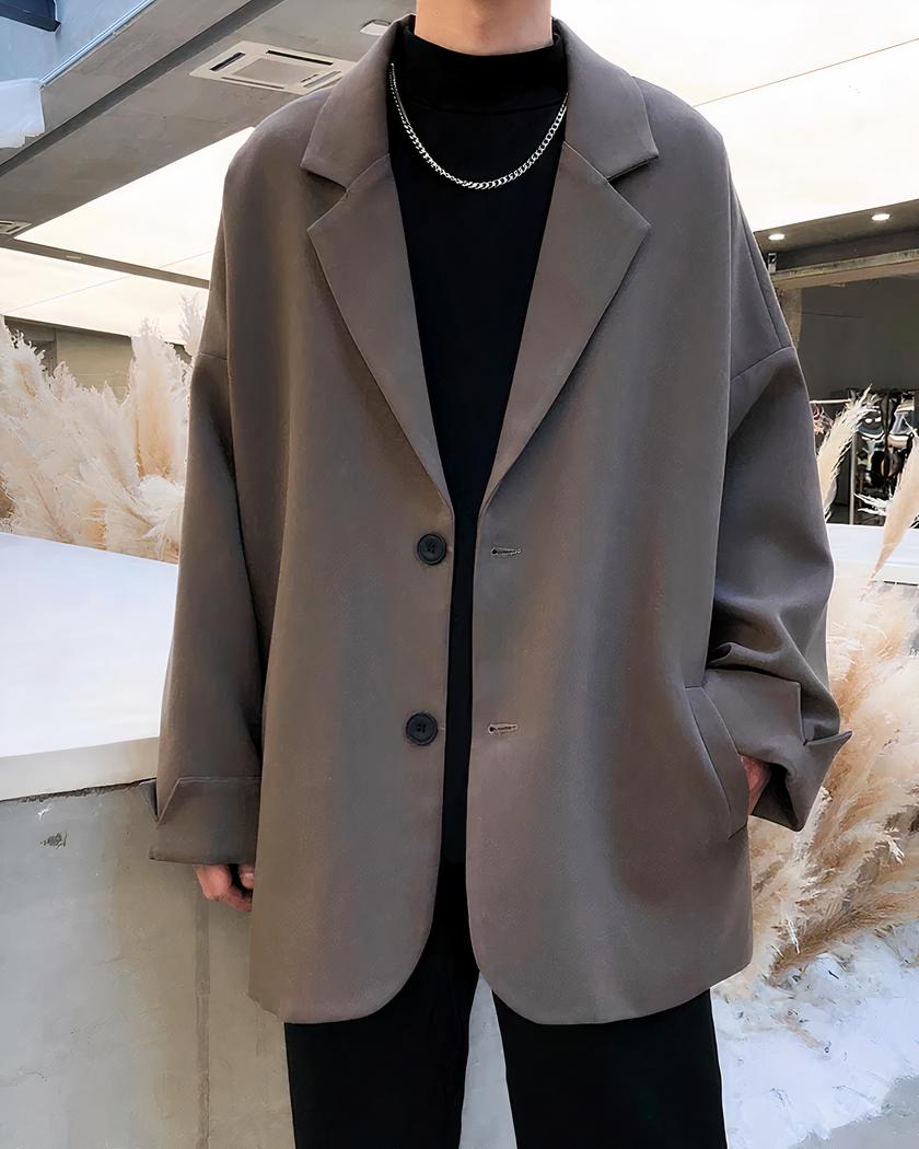 Blazer oversize homme manches à revers Photo d'un homme debout dans une grande pièce devant un meiuble blanc et des branches de feuillage beige portant un blazer oversize marron sur un pull col roulé noir et un pantalon noir large main gauche dans la poche du blazer à deux boutons noirs
