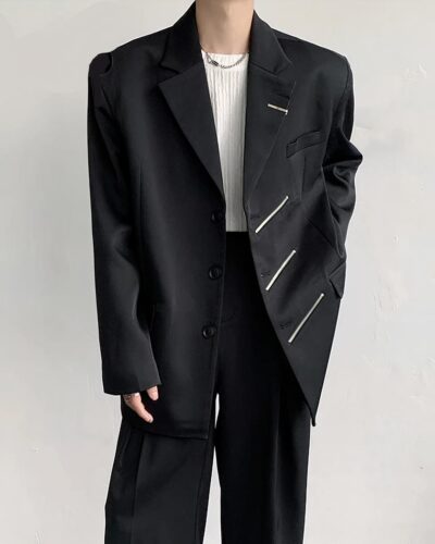 Photo d'un homme debout portant un blazer oversize noir orné d'une barrette au revers gauche et de trois fermetures éclair ou zips en oblique également à gauche, un pantalon noir à pinces et un teeshirt blanc col rond, le tout sur un fond blanc cassé