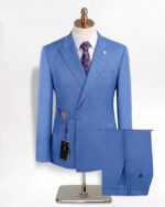 Photo d'un ensemble blazer croisé bleu ciel à fermeture par boucle d'acier dorée présenté sur un mannequin à pied argenté, une chemise blanche et une cravate en dessous, puis le pantalon plié sur un support horizontal, le tout sur un fond blanc