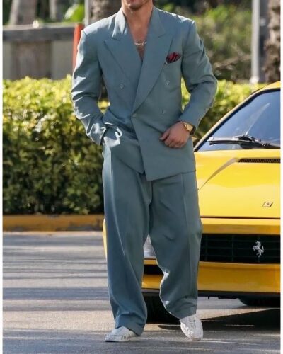 Photo d'un homme marchant devant une Ferrari jaune et portant un ensemble blazer croisé gris à pantalon large avec un epochette rouge dans la poche poitrine et un collier à grosses mailles argenté et des mocassins blancs