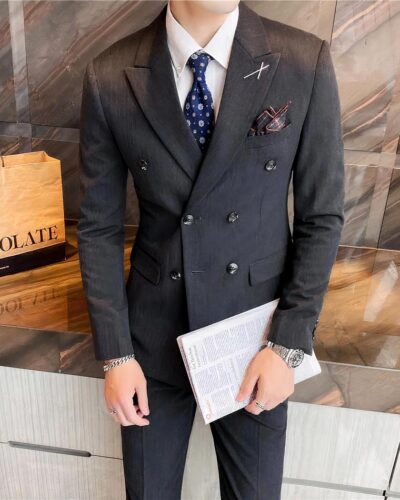 Photo d'un homme debout devant une vitre portant un ensemble blazer croisé 3 pièces à 6 boutons gris foncé un insigne représentant un X sur le revers gauche, avec une chemise blanche et une cravate gris foncé une pochette mordorée dans la poche poitrine dans la main gauche un fascicule