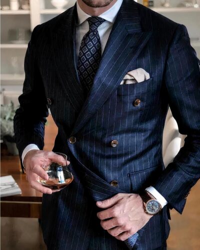 Photo d'un homme portant un ensemble blazer pantalon bleu marine rayé blanc croisé avec cravate à losanges bleus et blancs sur chemise blanche et une pochette gris clair dans la poche poitrine, une montre au poignet gauche, un verre d'alcool dans la main droite dans une pièce à étagères blanches au fond et une table à manger en bois