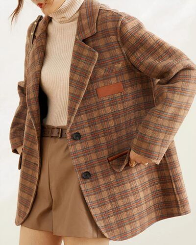 On voit une femme qui porte un pull col roulé beige et un short marron, ceinturé, associé à un manteau blazer en laine marron à carreaux, effet vintage