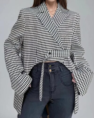 On voit une femme brune qui porte un manteau type coupe-vent, coupe blazer, motif pied-de-poule asymétrique court devant et long derrière