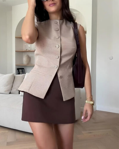Femme brune dans son salon portant un gilet blazer à petit carreaux avec une jupe couleur chocolat et un sac à main