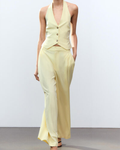 Femme portant un ensemble blazer pantalon femme jaune devant un mur blanc uni et sur un sol gris